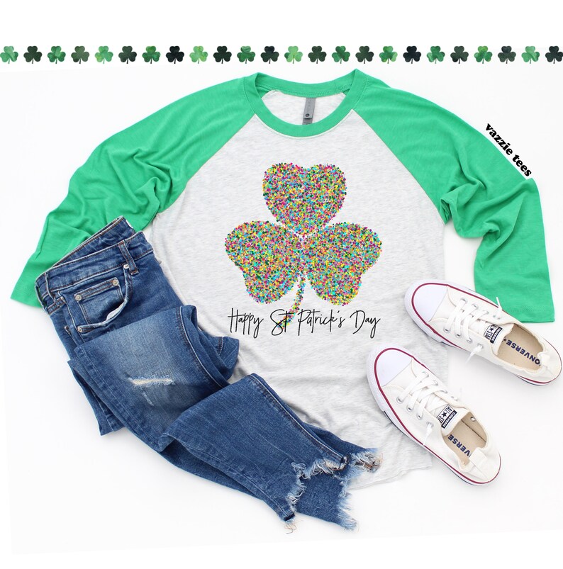 K&ouml;nnte beinhalten: Ein hellgraues und gr&uuml;nes Raglan-Shirt mit einem bunten Kleeblatt-Design und dem Text "Happy St. Patrick's Day".