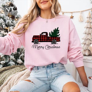 May include: A pink sweatshirt with a red and black plaid truck carrying a green Christmas tree. The text "Merry Christmas" is printed below the truck.