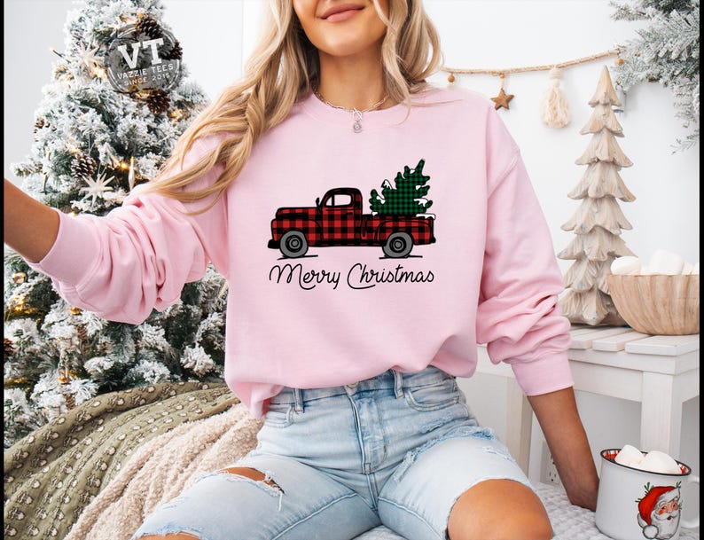 May include: A pink sweatshirt with a red and black plaid truck carrying a green Christmas tree. The text "Merry Christmas" is printed below the truck.