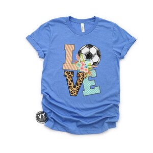 May include: A blue t-shirt with the word "LOVE" spelled out in colorful patterned letters. A soccer ball is on top of the "O" with flowers around it.