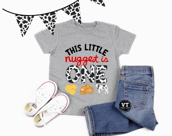 Könnte beinhalten: Ein graues T-Shirt mit einem Kuhmuster, auf dem "This little nugget is one" steht. Das Shirt wird mit einer blauen Jeans, weißen Turnschuhen und einem Kuhmusterbanner kombiniert.
