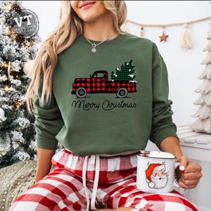 May include: A green sweatshirt with a red and black plaid truck carrying a green Christmas tree. The text "Merry Christmas" is printed below the truck. The model is wearing red and black plaid pajama pants and holding a white mug with a Santa Claus illustration.