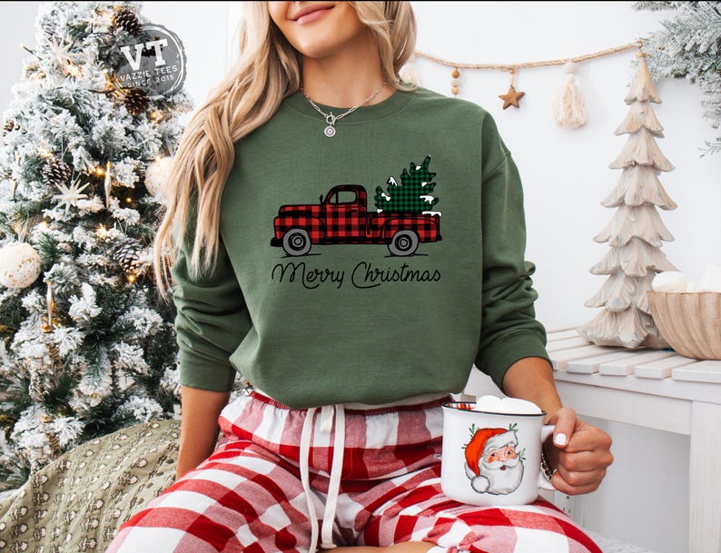 May include: A green sweatshirt with a red and black plaid truck carrying a green Christmas tree. The text "Merry Christmas" is printed below the truck. The model is wearing red and black plaid pajama pants and holding a white mug with a Santa Claus illustration.