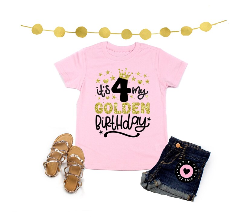 May include: A pink t-shirt with the text "it's my 4 GOLDEN BIRTHDAY" in gold glitter and black lettering. The image also includes denim shorts, gold sandals, and a gold star garland.
