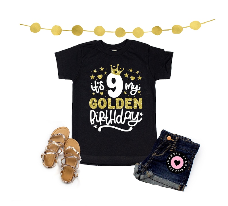 May include: Black t-shirt with gold glitter text that reads "it's 9 my GOLDEN BIRTHDAY" with a crown and stars. The image also shows denim shorts, gold sandals, and a gold garland.