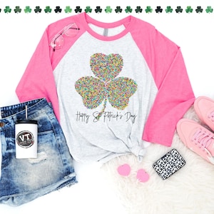 K&ouml;nnte beinhalten: Ein wei&szlig;es und pinkfarbenes Baseball-Shirt mit einem bunten Kleeblatt-Design und dem Text "Happy St. Patrick's Day".