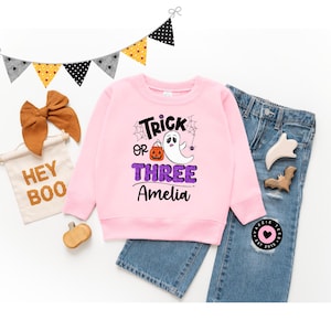 May include: A pink Halloween-themed sweatshirt with the text "Trick or Three" and a ghost graphic, paired with blue jeans. A banner reads "Hey Boo" and a brown bow and pumpkin are also included.