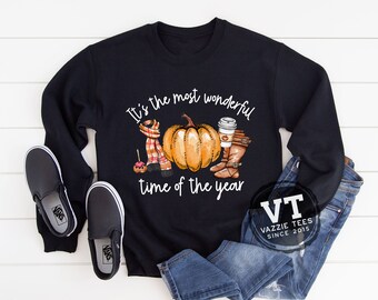 May include: Black sweatshirt with the text "It's the most wonderful time of the year." The graphic includes a pumpkin, scarf, coffee cup, boots, and an apple. Black slip-on shoes and blue jeans are also in the image.