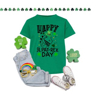 May include: A green t-shirt with the text "HAPPY St. PAT-REX DAY" and a cartoon dinosaur wearing a leprechaun hat. The image includes light blue jeans, white trainers, and St. Patrick's Day themed accessories like a rainbow and shamrock decorations.