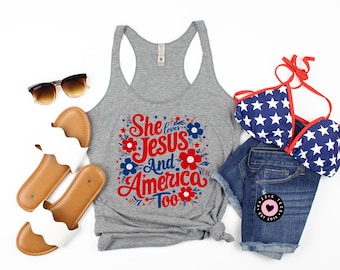 May include: A gray tank top with the text "She loves Jesus and America Too" in red, blue, and white lettering with floral accents. The tank top is laying on a white surface with a pair of brown sandals, a pair of white sandals, and a blue and white star patterned bikini top.