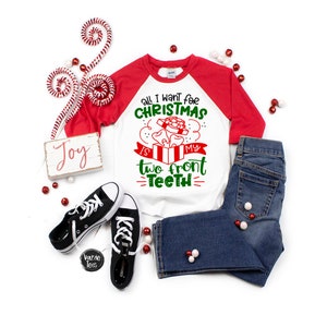 May include: A white and red raglan-style Christmas shirt with the text "All I want for Christmas is my two front teeth." The shirt is paired with blue jeans and black sneakers. Decorative candy canes and ornaments complete the festive arrangement.