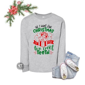 May include: A gray long-sleeved sweatshirt with a Christmas-themed design. The text reads "All I want for Christmas is my two front teeth". The design features a red and white gift box with two teeth inside.