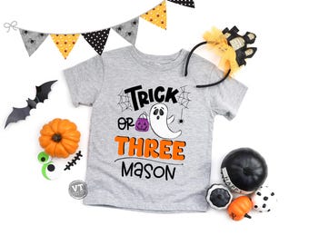 May include: A gray t-shirt with the text "Trick or Three Mason" and a ghost holding a pumpkin. The shirt is surrounded by Halloween decorations, including a pumpkin, a bat, and a spider.