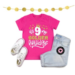 May include: A hot pink t-shirt with the text "It's 9 my Golden Birthday" in white and gold glitter. The shirt is paired with light wash denim shorts and white sneakers. A gold garland is at the top.