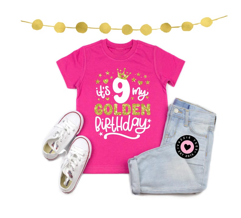 May include: A hot pink t-shirt with the text "It's 9 my Golden Birthday" in white and gold glitter. The shirt is paired with light wash denim shorts and white sneakers. A gold garland is at the top.