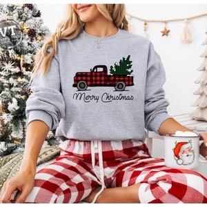 May include: A gray sweatshirt with a red and black plaid truck carrying a green Christmas tree. The truck has the words "Merry Christmas" printed below it.