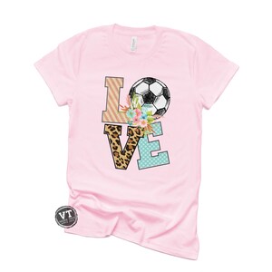 May include: Pink t-shirt with a graphic of the word "LOVE" with a soccer ball and floral accents. The letters are decorated with different patterns, including leopard print, stripes, and polka dots.