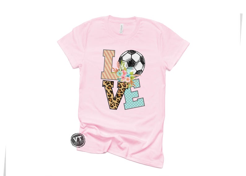May include: Pink t-shirt with a graphic of the word "LOVE" with a soccer ball and floral accents. The letters are decorated with different patterns, including leopard print, stripes, and polka dots.