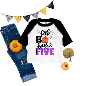 May include: A white and black raglan shirt with the text "fab BOO lous & FIVE" and a pumpkin and ghost graphic. The image also includes a pair of blue jeans, decorative pumpkins, and a Halloween banner.