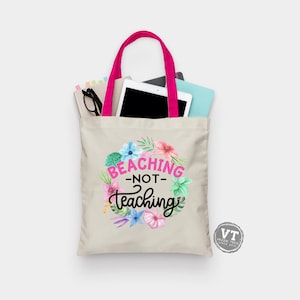 May include: A beige tote bag with pink handles and a floral design. The bag has the text "Beaching - Not - Teaching" in black lettering.