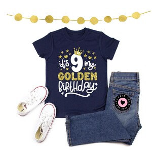 May include: Navy blue t-shirt with the text "it's 9 my GOLDEN Birthday" in gold and white, paired with denim jeans and white sneakers. A gold garland is at the top. The jeans have a pink heart patch.