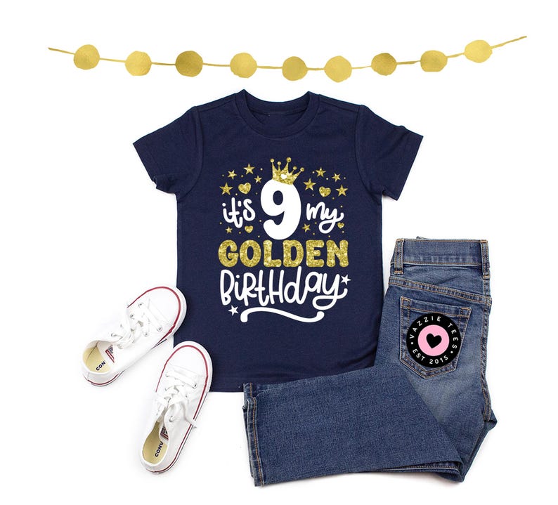 May include: Navy blue t-shirt with the text "it's 9 my GOLDEN Birthday" in gold and white, paired with denim jeans and white sneakers. A gold garland is at the top. The jeans have a pink heart patch.