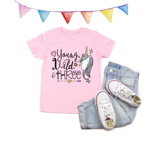 May include: Pink t-shirt with a unicorn graphic and the text "Young, Wild & Three". The shirt is paired with light blue jeans and white sneakers.