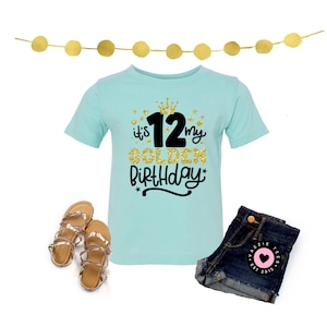 May include: A light blue t-shirt with the text "it's my GOLDEN BIRTHDAY 12". The shirt is paired with denim shorts and gold sandals. A gold garland is at the top of the image. The shorts have a pink heart patch.