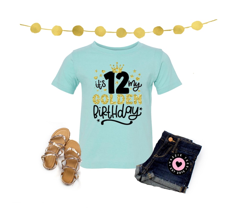 May include: A light blue t-shirt with the text "it's my GOLDEN BIRTHDAY 12". The shirt is paired with denim shorts and gold sandals. A gold garland is at the top of the image. The shorts have a pink heart patch.