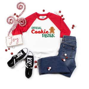 May include: A white and red baseball style shirt with the text "OFFICIAL Cookie TASTER" in green and red lettering with a gingerbread man graphic. The shirt is laying on a white surface with red and white candy canes, red and white beads, and a wooden sign that says "Joy".
