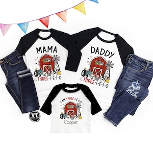 Peut inclure: Un ensemble de trois t-shirts assortis représentant une grange rouge, un tracteur et le texte "I am Three-I-E-I-O" sur un t-shirt blanc, "Mama of the Three-I-E-I-O" sur un t-shirt noir et blanc, et "Daddy of the Three-I-E-I-O" sur un t-shirt noir et blanc.