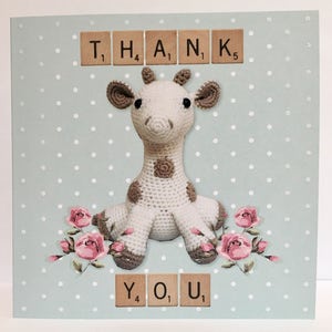 May include: A light blue card with white polka dots features a crocheted giraffe toy sitting on a bed of pink roses. The card says "THANK YOU" in Scrabble tiles.