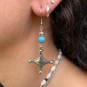 May include: Silver earrings with a turquoise bead and a star-shaped charm. The earrings are hanging from a person's ear.
