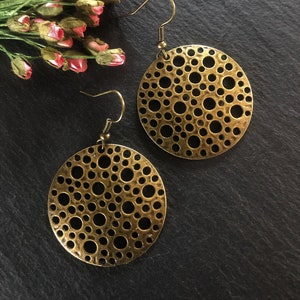 May include: A pair of gold-toned earrings with a circular design featuring a pattern of small, evenly spaced holes. The earrings have a simple hook closure.