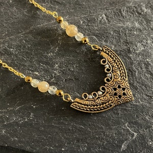 May include: A gold necklace with a large, ornate pendant featuring a crescent moon design. The pendant is decorated with intricate details and small, clear rhinestones. The necklace is strung with small, light brown and clear beads.