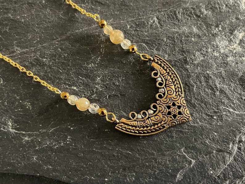 May include: A gold necklace with a large, ornate pendant featuring a crescent moon design. The pendant is decorated with intricate details and small, clear rhinestones. The necklace is strung with small, light brown and clear beads.