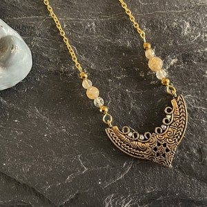 May include: A gold chain necklace with a gold pendant featuring an intricate design. The pendant is shaped like a crescent moon and has small clear crystals embedded in it. The chain is adorned with small, light brown beads.