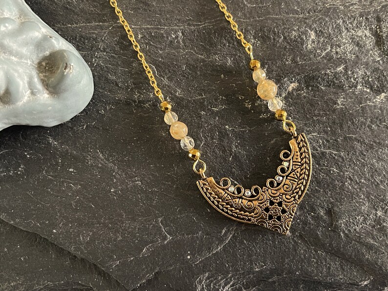 May include: A gold chain necklace with a gold pendant featuring an intricate design. The pendant is shaped like a crescent moon and has small clear crystals embedded in it. The chain is adorned with small, light brown beads.