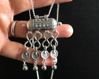 May include: A silver necklace with a detailed pendant featuring a cylindrical charm with intricate designs and a dangling chain with circular charms and small bells.