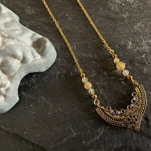 May include: A gold chain necklace with a crescent moon pendant. The pendant is decorated with intricate details and has small clear crystals. The chain is adorned with small, round, light brown and clear beads.