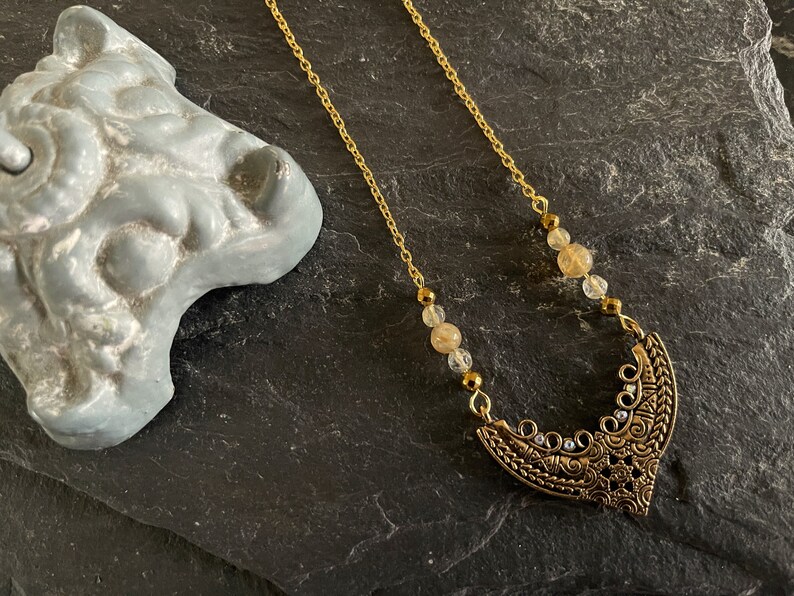 May include: A gold chain necklace with a crescent moon pendant. The pendant is decorated with intricate details and has small clear crystals. The chain is adorned with small, round, light brown and clear beads.