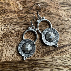 May include: Silver earrings with a circular design and a textured, faceted stone in the center. The earrings have a hoop design with a small bead detail.
