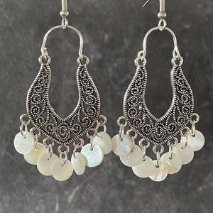May include: A pair of silver-toned dangle earrings featuring an ornate, filigree-style metal arch at the top. Suspended from the bottom of each metal arch are multiple small, round, iridescent white shell discs that hang freely as charms.