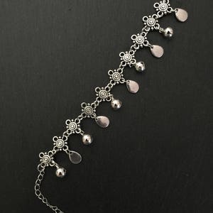 May include: Silver anklet with a chain and decorative charms. The charms are small, round, and have a floral design. The anklet has a dangling teardrop-shaped charm with a bell at the end.