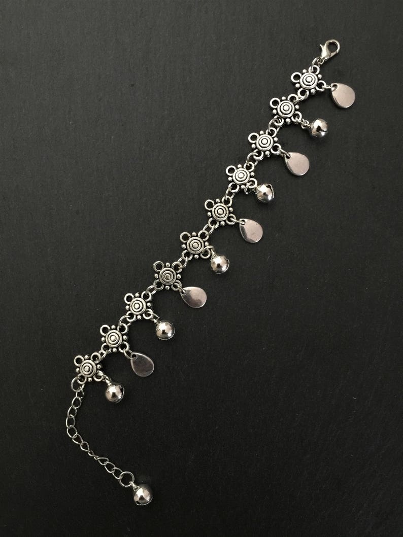 May include: Silver anklet with a chain and decorative charms. The charms are small, round, and have a floral design. The anklet has a dangling teardrop-shaped charm with a bell at the end.