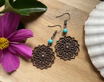 Puede incluir: Un par de pendientes de filigrana de color cobre con cuentas turquesas. Los pendientes tienen un diseño floral y se exhiben sobre una superficie de madera, junto a una flor morada y una concha blanca.