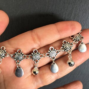May include: Silver chain anklet with decorative charms and small bells. The charms are round with a circle design and the bells are teardrop shaped.