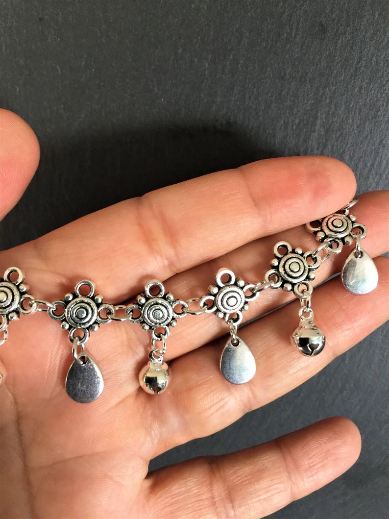 May include: Silver chain anklet with decorative charms and small bells. The charms are round with a circle design and the bells are teardrop shaped.