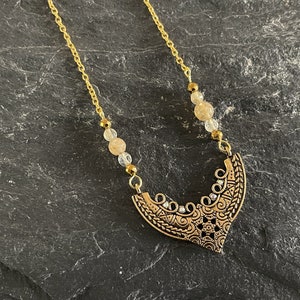 May include: A gold chain necklace with a gold pendant featuring an intricate design. The pendant is shaped like a crescent moon and has small clear stones embedded in it. The necklace is accented with small, light brown beads.