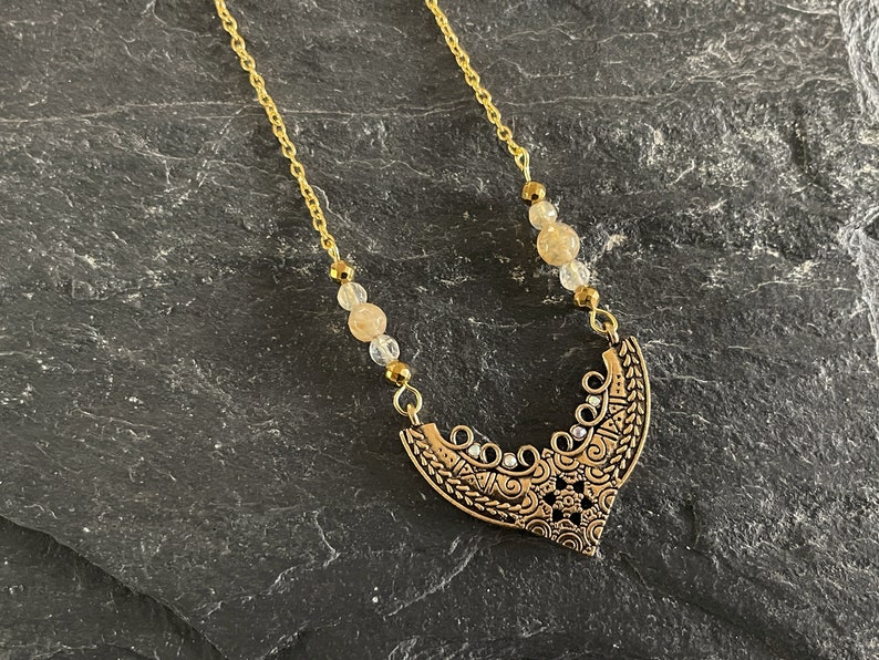 May include: A gold chain necklace with a gold pendant featuring an intricate design. The pendant is shaped like a crescent moon and has small clear stones embedded in it. The necklace is accented with small, light brown beads.